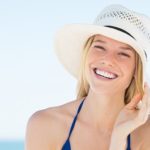 Portrait of smiling woman on beach wearing blue bikini and straw hat.