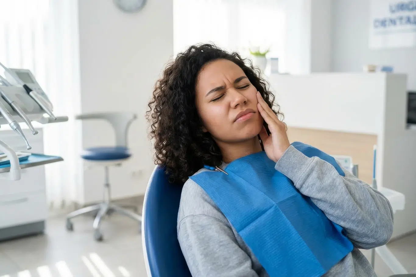 A young woman with curly hair sitting in a dental chair wearing a blue bib grimacing in pain and holding her cheek